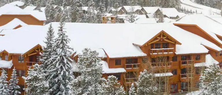 Vista aérea de um complexo de chalés em uma área de montanha, rodeado por árvores nevadas e pistas de esqui.