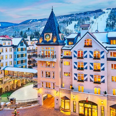 Vista de hotel com arquitetura alpina iluminada, montanhas nevadas ao fundo.