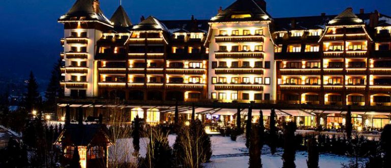 Fachada iluminada do The Alpina Gstaad, hotel luxuoso cercado por neve à noite.