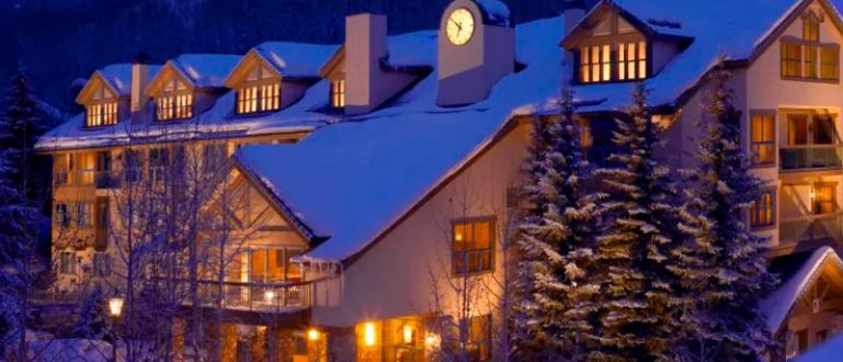 Hotel iluminado ao anoitecer, com neve no telhado e montanhas ao fundo.