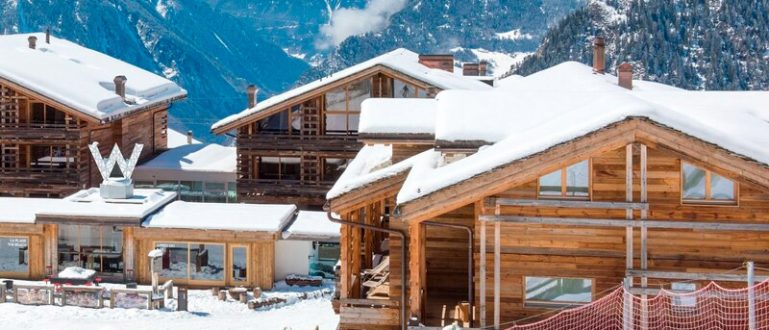 Chalés de madeira na neve com esquiadores nas proximidades, montanhas ao fundo.